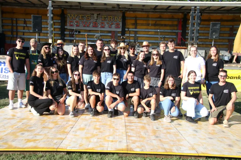 Eine Gruppe von Menschen in schwarzen T-Shirts mit dem Text 'GVP' posiert für ein Foto auf einer Bühne mit Holzboden. Dahinter befindet sich eine gelbe Wand mit dem Text 'Western Cowboys' und Musikinstrumenten.