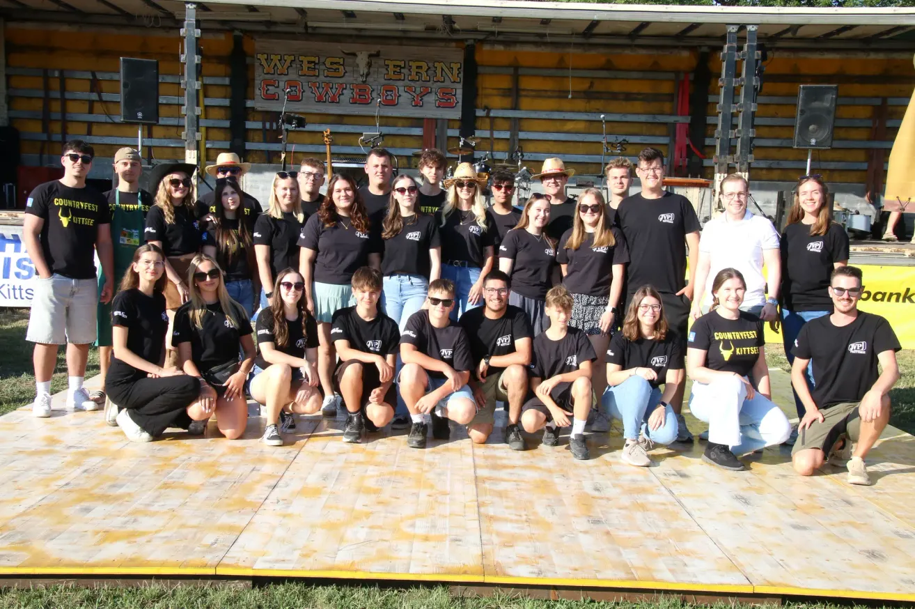 Eine Gruppe von Menschen in schwarzen T-Shirts mit dem Text 'GVP' posiert für ein Foto auf einer Bühne mit Holzboden. Dahinter befindet sich eine gelbe Wand mit dem Text 'Western Cowboys' und Musikinstrumenten.