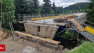 Eine Baustelle mit einem großen Betonpfeiler und Metallgerüsten, Holzbrettern und Metallzäunen. Bäume und ein Fluss sind im Hintergrund.