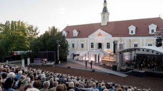 Eine klassische Musikaufführung findet vor einem großen, historischen Gebäude mit rotem Dach und Turm statt. Die Bühne ist mit Lautsprechern und Beleuchtungsequipment ausgestattet. Zwei Künstler stehen auf der Bühne, und ein großes Publikum sitzt davor.