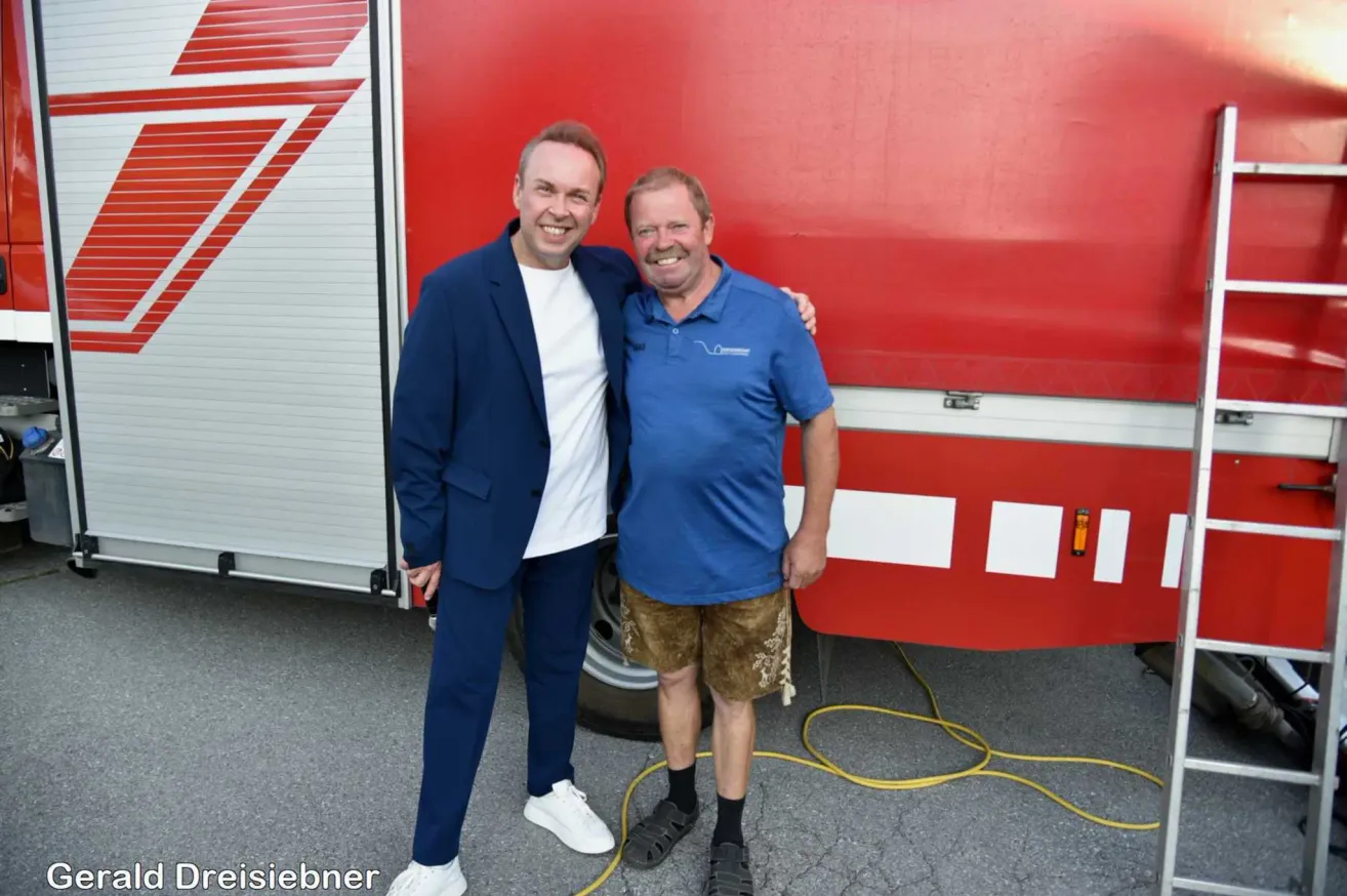 Zwei Männer stehen vor einem roten Feuerwehrauto, einer trägt einen blauen Anzug und der andere ein blaues Poloshirt und Shorts. Sie lächeln beide.