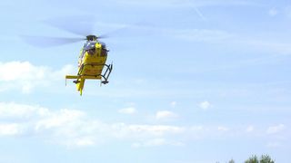 Ein gelber Hubschrauber mit der Registrierung OE-XLA fliegt in einem klaren blauen Himmel mit verstreuten Wolken.