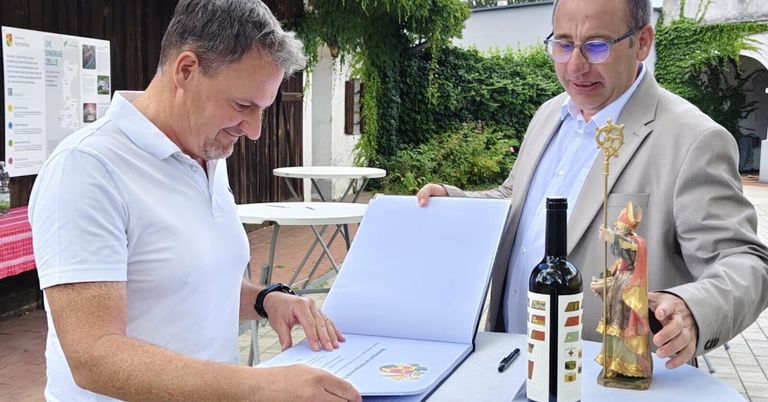 Zwei Männer in formeller Kleidung stehen draußen, einer hält ein offenes Buch und der andere eine Flasche Wein. Sie lächeln und scheinen ein Dokument zu unterzeichnen.