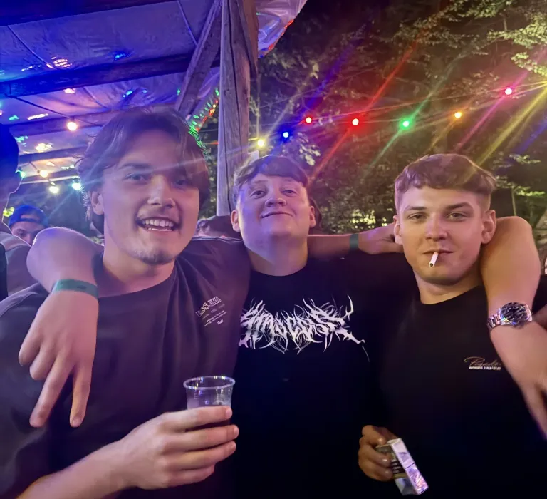 Three young men stand together at a party, one holding a drink and another smoking a cigarette. They are under a canopy with colorful lights.