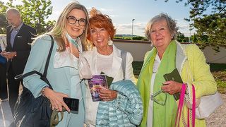 Drei Frauen stehen zusammen in einer Außenumgebung, lächeln und halten Bücher. Die Frau links trägt eine Brille und einen blauen Mantel, die mittlere eine weiße Jacke und eine blaue Jacke und die rechte trägt einen grünen Schal und einen gelben Mantel. Ein Mann steht hinter ihnen.
