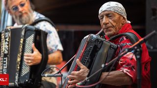 Ein älterer Mann spielt Akkordeon, während ein anderer Mann mit Bart ein weiteres Akkordeon im Hintergrund spielt. Beide tragen Bandanas.