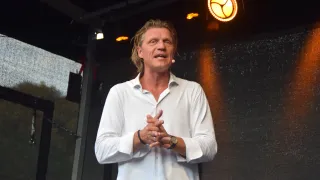 Ein Mann mit langen Haaren und einem weißen Hemd steht auf einer Bühne und hält möglicherweise eine Rede. Er hat die Hände zusammengepresst und trägt eine Uhr und einen Ring. Hinter ihm befindet sich eine dunkle Wand, die von einem gelben Licht beleuchtet wird.