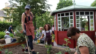 Eine Gruppe von Frauen in Schürzen pflegt einen Garten mit einem roten Pavillon im Hintergrund. Sie pflanzen Topfpflanzen in den Boden.