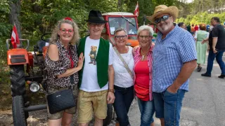 Fünf Menschen stehen zusammen vor einem Traktor. Sie alle lächeln und scheinen für ein Foto zu posieren. Der Mann in der Mitte trägt eine grüne Weste mit einem Bären darauf.