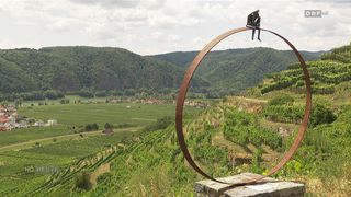 Eine große rostige Metallring-Skulptur steht auf einem Hügel mit Blick auf eine Weinberglandschaft. Eine Statue eines Paares sitzt auf dem Ring.