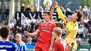 Fußballspieler in roten und gelben Trikots befinden sich in einem heftigen Spiel, wobei ein Spieler versucht, den Ball zu köpfen, während ein anderer verteidigt. Das Publikum ist im Hintergrund verschwommen.