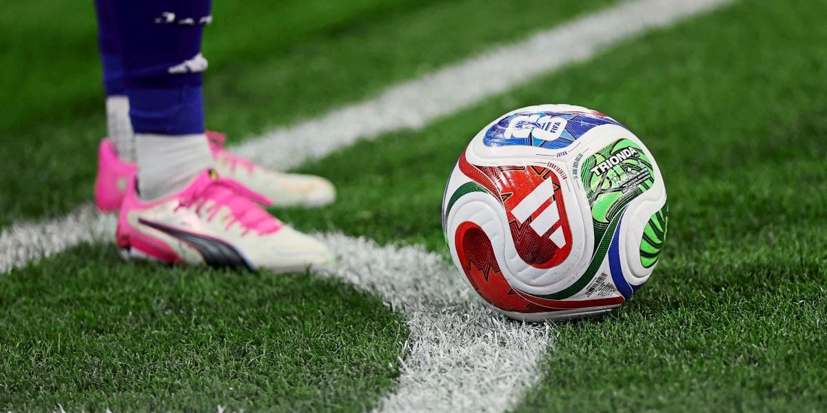 Ein Nahaufnahme eines Fußballs mit einem bunten Design auf einem grünen Feld, neben dem Fuß eines Spielers in pinkfarbenen Stollenschuhen.