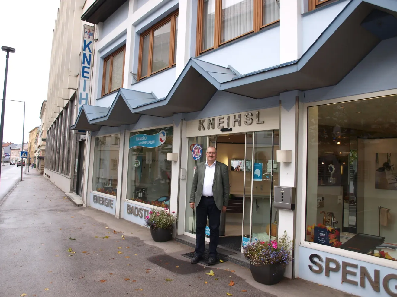 Ein Mann steht vor einem blau-weißen Gebäude mit dem Wort Kneihsl darauf. Das Gebäude hat Glastüren und Fenster.