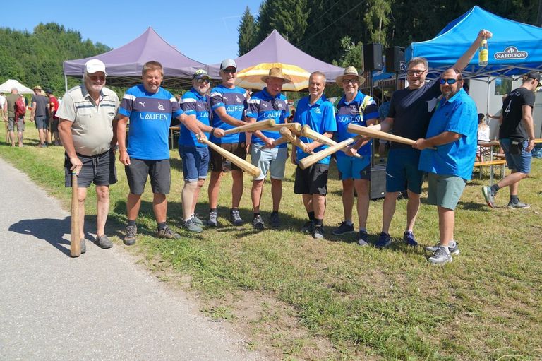 Eine Gruppe von Männern in blauen Shirts mit dem Wort Laufen darauf halten Holzschläger in einer Outdoor-Umgebung. Sie stehen auf einer Rasenfläche mit Zelten im Hintergrund.