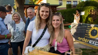 Zwei Frauen lächeln für ein Foto bei einer Outdoor-Veranstaltung mit Getränken auf einem Tisch und Leuten im Hintergrund.