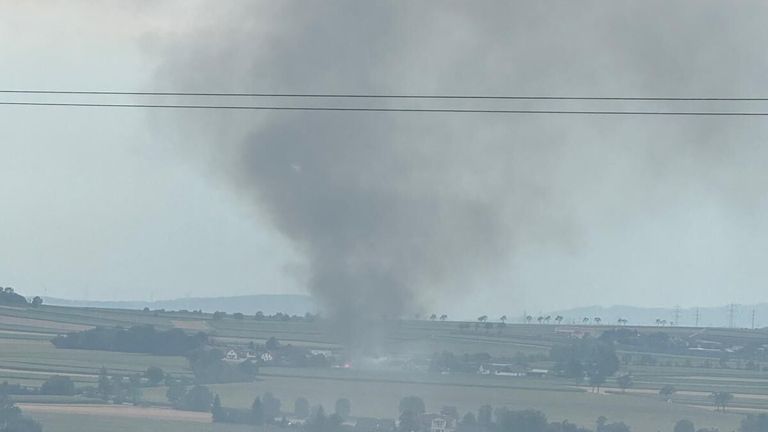 Ein großer Rauchschwaden steigt aus einem ländlichen Gebiet auf, mit einem in der Ferne sichtbaren Feuer zwischen Bäumen und Gebäuden.