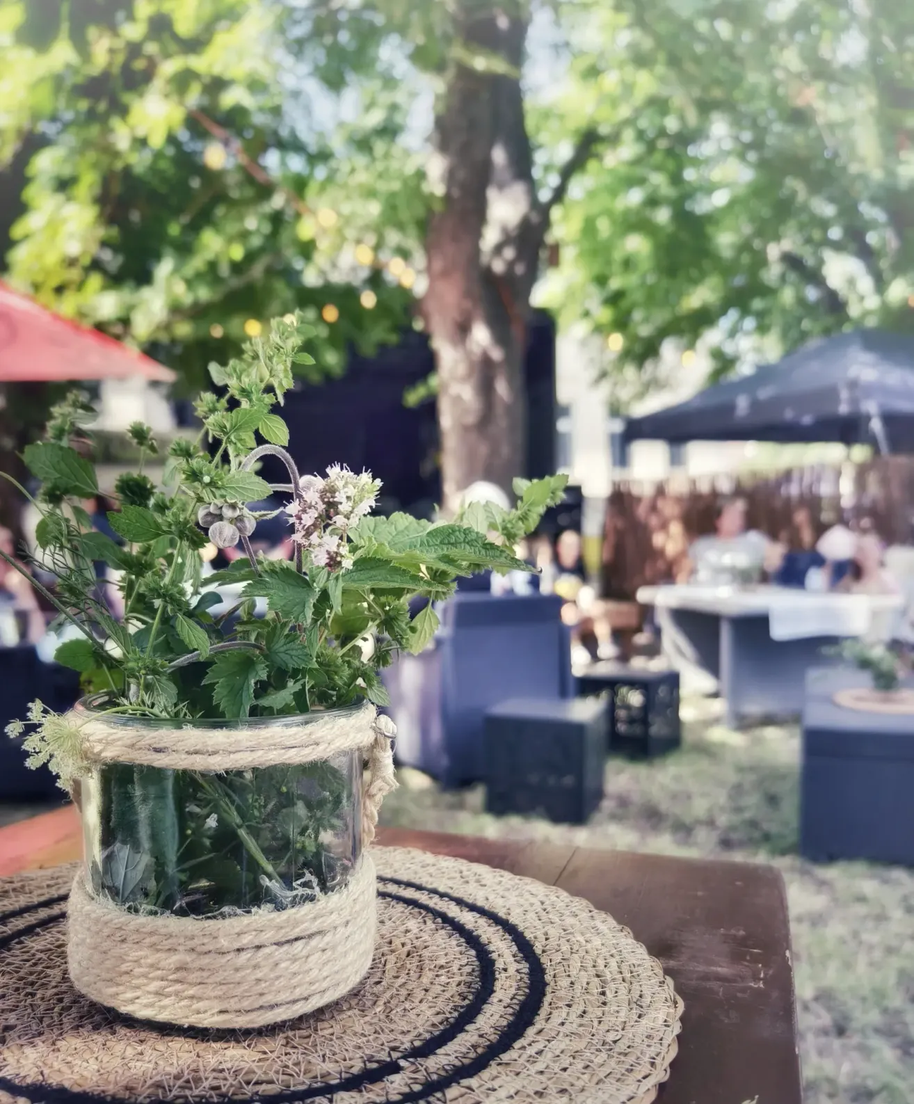 Ein rustikales Glasvase mit einem umwickelten Seil, gefüllt mit Kräutern und Blumen, auf einem Tisch in einer Gartenumgebung im Freien.