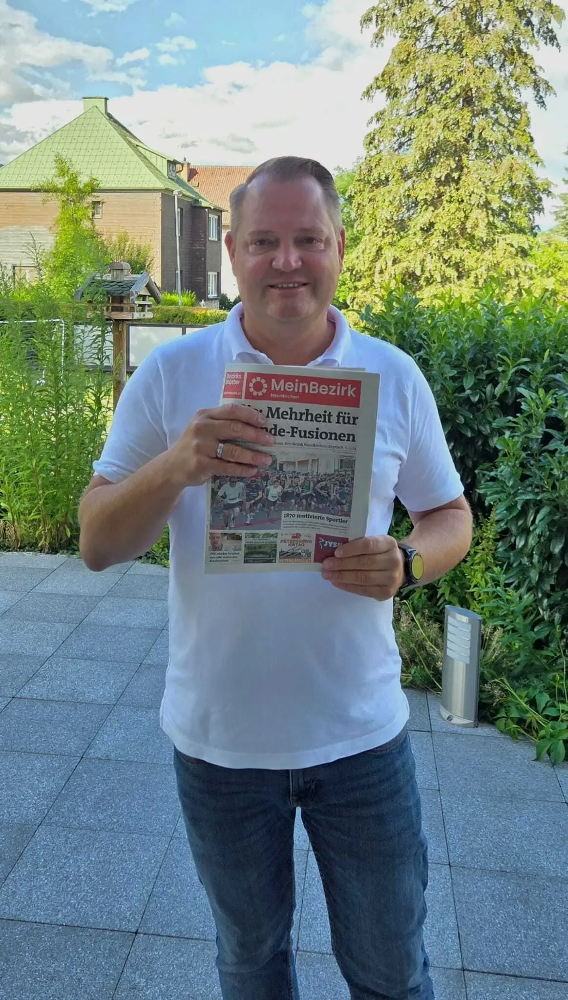 Ein Mann in einem weißen Hemd hält eine Zeitung mit der Überschrift 'Mehrheit für die Fusionen'. Er steht draußen mit Grünpflanzen und einem Gebäude im Hintergrund.