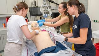 Drei Frauen in einem medizinischen Umfeld führen eine Herz-Lungen-Wiederbelebung an einem Mannequin durch. Die Frau auf der linken Seite hält den Kopf des Mannequins, während die Frau in der Mitte einen Defibrillator benutzt. Die Frau auf der rechten Seite beobachtet.