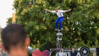 Ein Mann führt einen Balanceakt auf einem hohen Stapel silberner Rohre im Freien aus.