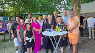 Eine Gruppe von Menschen, einschließlich Erwachsener und Kindern, steht in einer Außenumgebung zusammen. Sie lächeln und posieren für ein Foto. Vor ihnen steht ein Tisch mit einer Flasche, einem Becher und einem Teller. Hinter ihnen befinden sich Bäume und ein Gebäude.