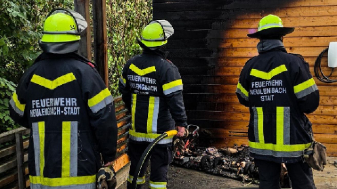 Drei Feuerwehrleute in Schutzkleidung stehen in der Nähe einer verbrannten Holzstruktur. Einer hält einen Schlauch, ein anderer schaut zu und der dritte schaut weg.