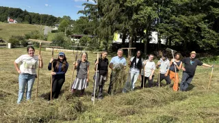 Eine Gruppe von Menschen auf einem Feld, mit Rechen in der Hand, lächeln für ein Foto. Bäume und ein Haus sind im Hintergrund.