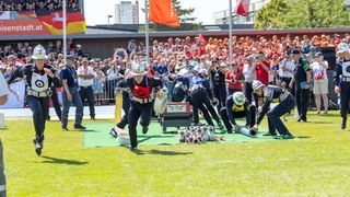 Eine Gruppe von Feuerwehrleuten in Uniform nimmt an einem Rennen auf einem Feld teil, während Zuschauer hinter einem Zaun zusehen.