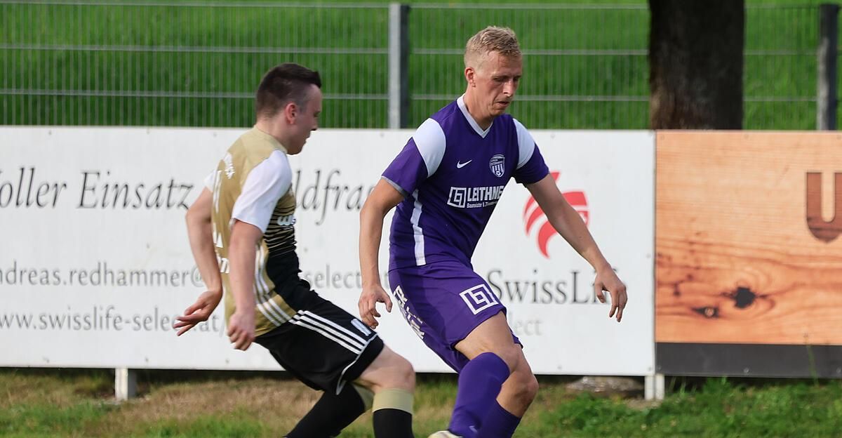 Zwei Fußballspieler rennen auf einem Feld. Ein Spieler trägt ein lila Trikot mit einem Logo auf der Brust. Der andere Spieler trägt ein gold-schwarzes Trikot. Hinter ihnen steht ein Zaun mit einem Banner.