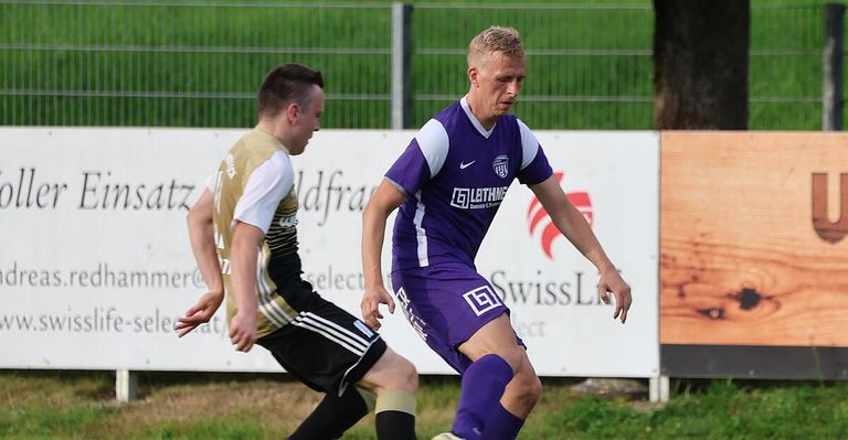 Zwei Fußballspieler rennen auf einem Feld. Ein Spieler trägt ein lila Trikot mit einem Logo auf der Brust. Der andere Spieler trägt ein gold-schwarzes Trikot. Hinter ihnen steht ein Zaun mit einem Banner.