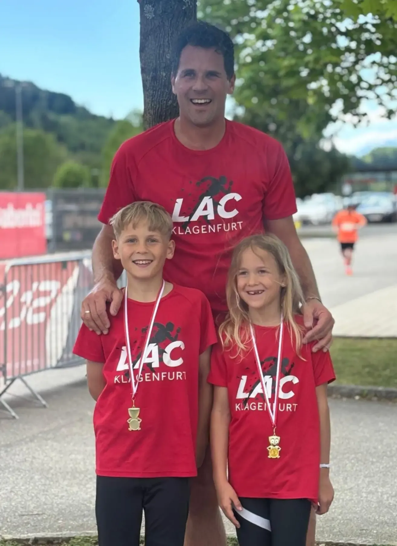 Ein Mann und zwei Kinder stehen zusammen und tragen rote T-Shirts mit dem Text 'LAC Klagenfurt' und halten Medaillen. Sie lächeln für ein Foto.