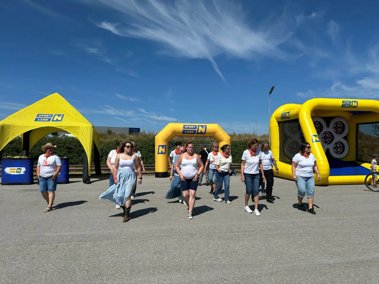 Eine Gruppe von Frauen in weißen Shirts und Jeans geht durch eine Outdoor-Veranstaltung mit aufblasbaren Strukturen und einem gelben Zelt.