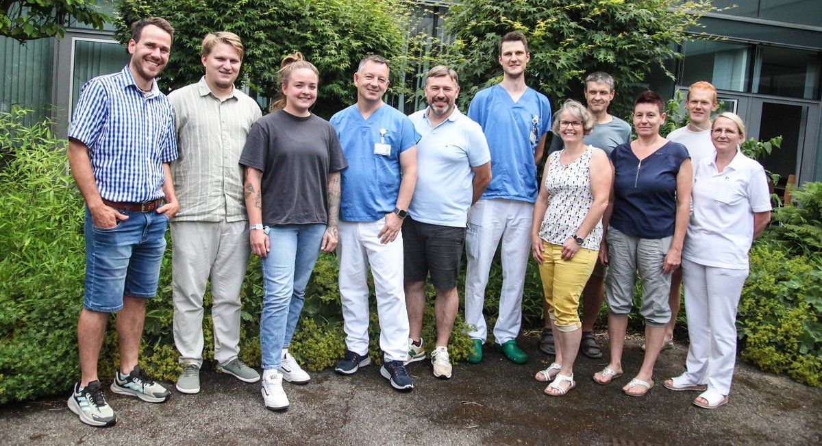 Eine Gruppe von Menschen, darunter Männer und Frauen, steht vor einem grünen Busch. Sie alle lächeln und scheinen für ein Foto zu posieren. Einige von ihnen tragen Turnschuhe, während andere Sandalen tragen.
