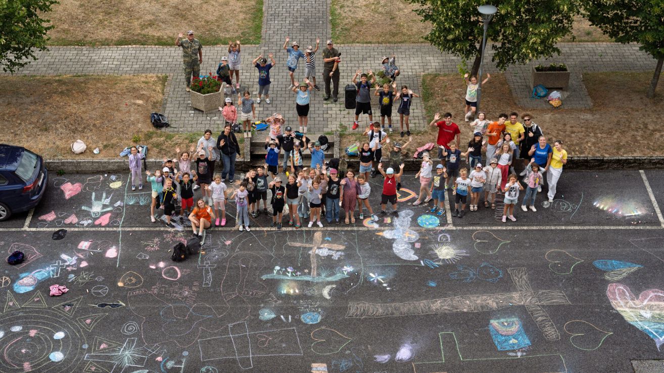Eine Gruppe von Kindern und Erwachsenen versammelt sich um eine Kreidezeichnung auf einem gepflasterten Bereich. Einige heben die Hände, während andere in der Nähe von Topfpflanzen und einem Baum stehen.