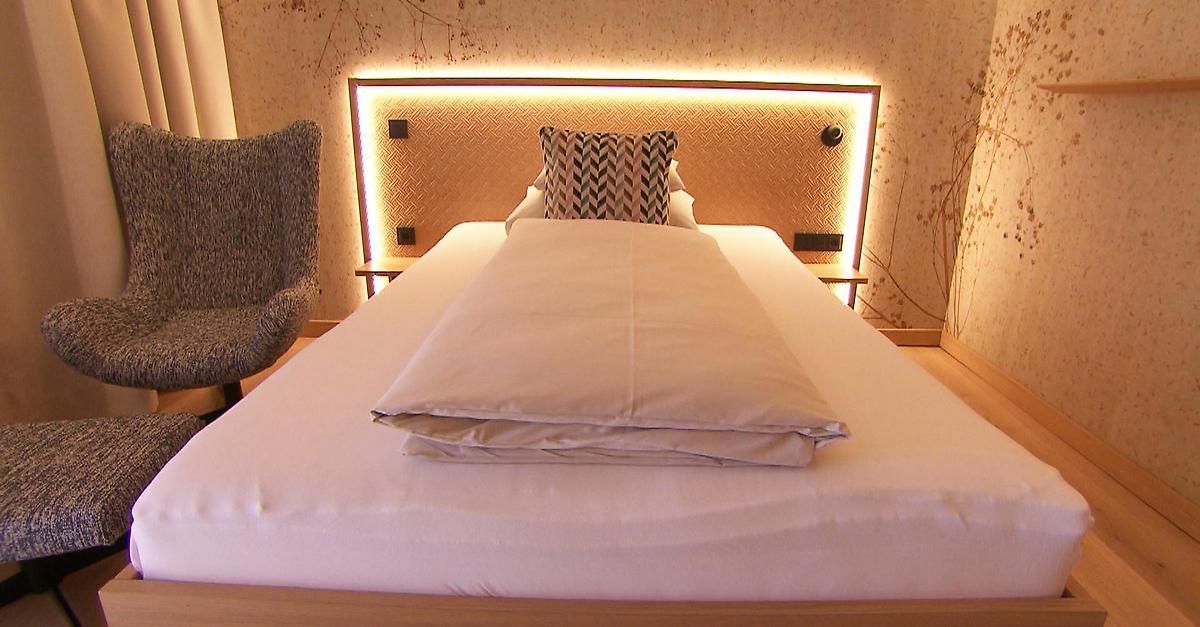 Ein ordentlich gemachtes Bett mit weißen Laken und einem gemusterten Kissen auf einem Holzbettrahmen, beleuchtet von warmen LED-Lichtern am Kopfteil.