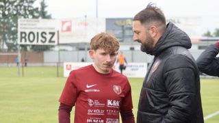 Ein junger Fußballspieler in einem roten Trikot mit verschiedenen Sponsorenlogos schaut einen Mann in einer schwarzen Jacke an, der mit ihm spricht. Sie stehen auf einem Fußballfeld mit einem verschwommenen Hintergrund.