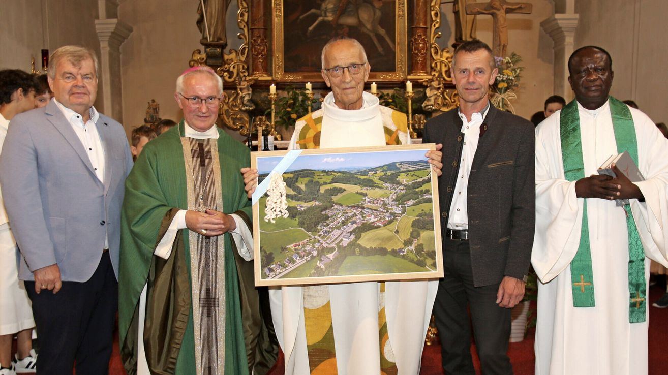 Drei Männer stehen in einer Kirche, einer hält ein gerahmtes Gemälde eines Dorfes. Der Mann in der Mitte trägt eine weiße Robe und eine Brille. Der Mann auf der linken Seite trägt eine grüne Robe mit einem Kreuz. Der Mann auf der rechten Seite trägt einen schwarzen Anzug.