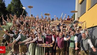 Eine große Gruppe von Menschen in traditioneller deutscher Kleidung, die Holzschilder mit Namen von Regionen halten, posiert für ein Foto vor einem Gebäude.