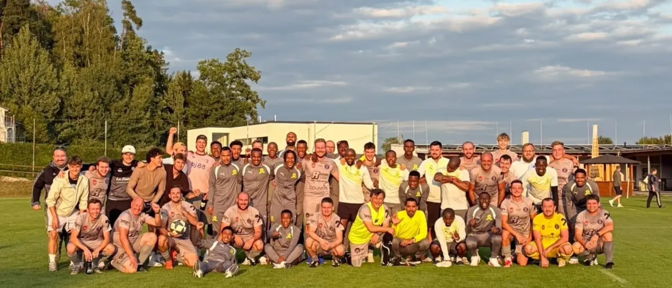 Eine Fußballmannschaft posiert für ein Gruppenfoto auf einem Rasenfeld mit einem Gebäude und einem Baum im Hintergrund.