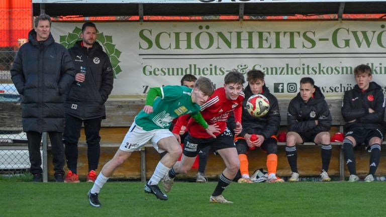 Zwei Fußballspieler in grünen und roten Trikots kämpfen um den Ball. Dahinter steht ein Mann in der Nähe einer Bank mit einem Schild, auf dem 'SCHÖNHEIT' steht.