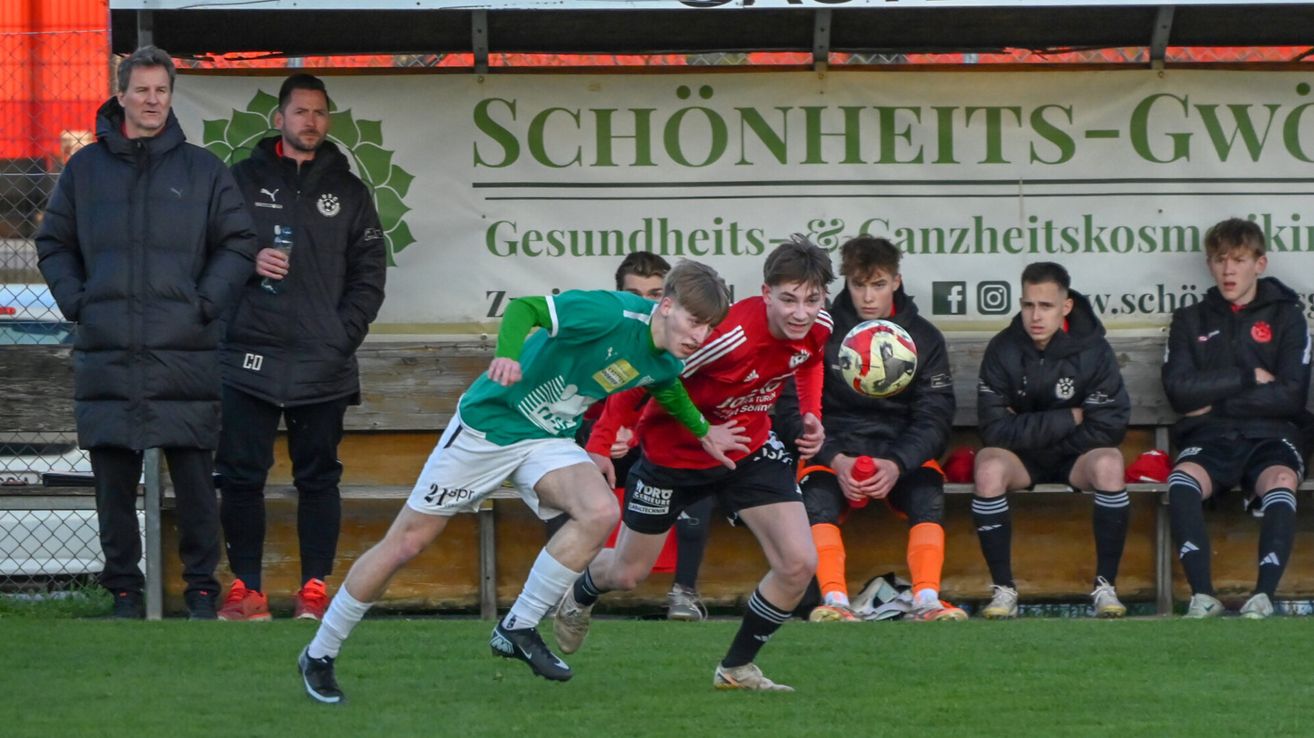 Zwei Fußballspieler in grünen und roten Trikots kämpfen um den Ball. Dahinter steht ein Mann in der Nähe einer Bank mit einem Schild, auf dem 'SCHÖNHEIT' steht.
