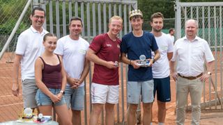 Fünf Personen stehen auf einem Tennisplatz, zwei von ihnen halten Pokale. Ein Mann trägt eine Krone und ein anderer hält einen Pokal. Hinter ihnen ist ein Metallzaun und Bäume zu sehen.
