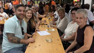 Eine Gruppe von Menschen sitzt an einem langen Holztisch im Freien und lächelt für ein Foto. Der Tisch ist mit Gläsern und Papieren bestückt. Im Hintergrund sitzen weitere Menschen unter Sonnenschirmen.