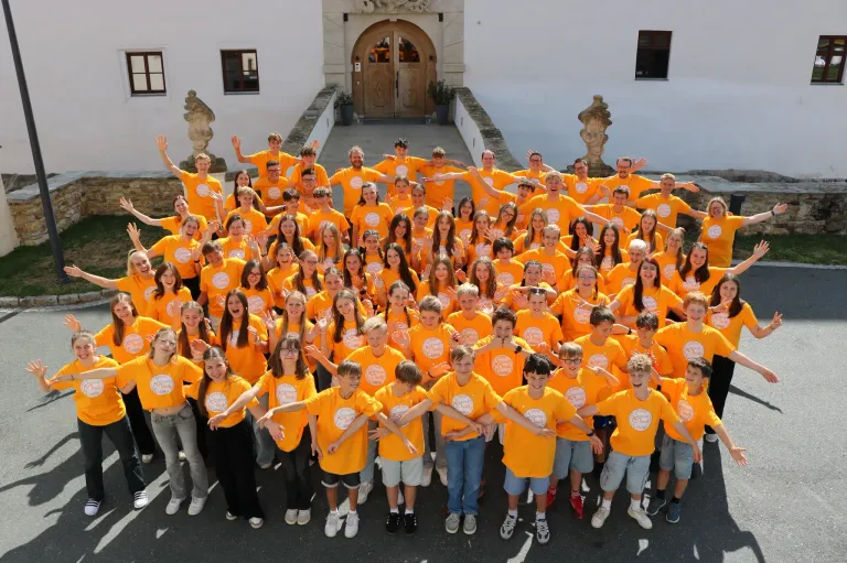 Eine große Gruppe junger Menschen in passenden orangefarbenen T-Shirts posiert für ein Foto vor einem Gebäude.