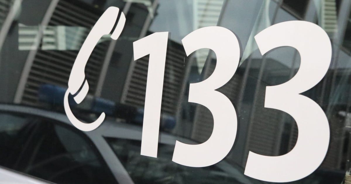 Eine Nahaufnahme eines weißen Notfallnummernschildes '133' auf einer Glasoberfläche mit einem unscharfen Hintergrund von Gebäuden und Fahrzeugen.