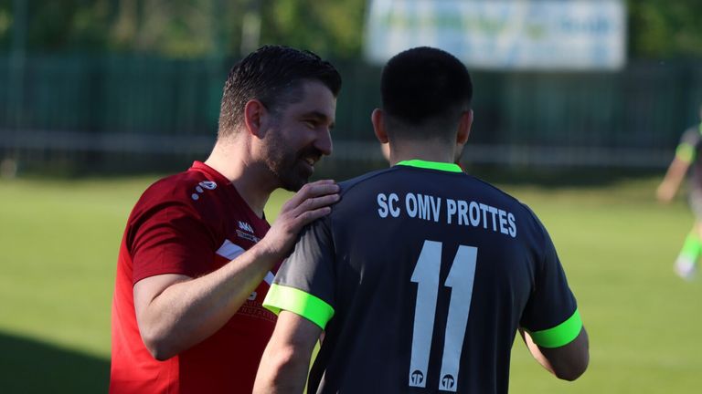 Zwei Männer in Sporttrikots, einer mit einem roten Shirt und der andere mit einem schwarzen Trikot mit der Nummer 11, stehen nah beieinander, wahrscheinlich auf einem Fußballfeld.