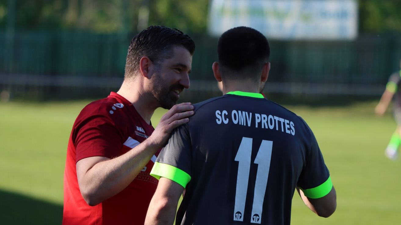 Zwei Männer in Sporttrikots, einer mit einem roten Shirt und der andere mit einem schwarzen Trikot mit der Nummer 11, stehen nah beieinander, wahrscheinlich auf einem Fußballfeld.