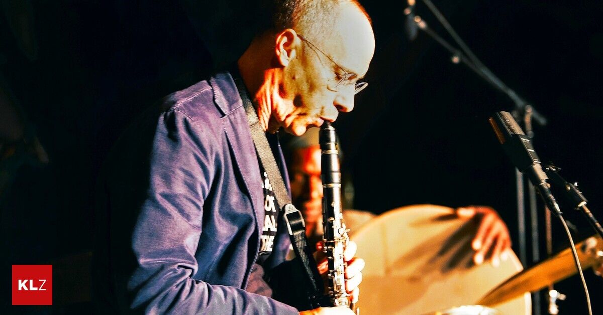 Ein Mann mit Brille und einem lila Hemd spielt Klarinette auf der Bühne, mit einem verschwommenen Hintergrund eines anderen Musikers, der eine Trommel spielt.