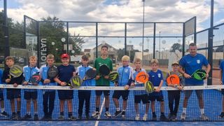 Eine Gruppe junger Jungen posiert für ein Foto auf einem Tennisplatz und hält Tennisschläger. Sie tragen Sportuniformen und Turnschuhe. Dahinter steht ein Zaun mit einem Netz, und in der Ferne sind Gebäude und Bäume zu sehen.