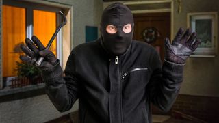 Eine Person in einer schwarzen Maske und Handschuhen hält einen Brecheisen und steht vor einem Haus.